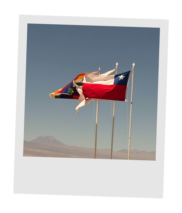 The Chilean flag flying beside the rainbow-checkered Wiphala flag of the Andean peoples, with a volcano on the desert horizon.