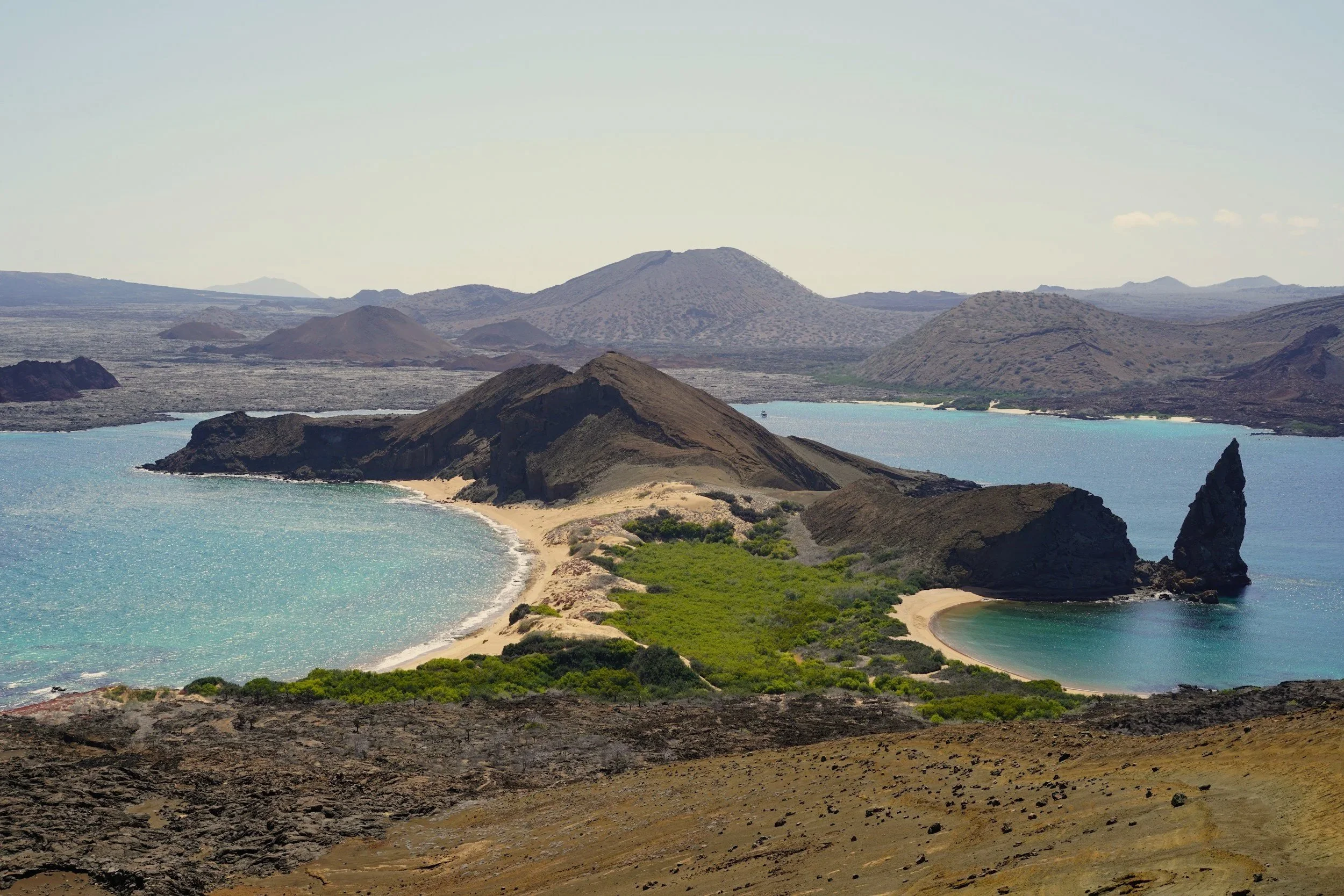 Aerial view of Bartolomé Island, with Pinnacle Rock rising beside a curved golden beach and two turquoise bays in the Galápagos.