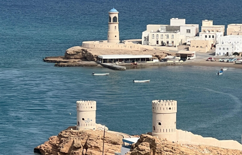 View of a coastal town with historic white buildings, a lighthouse on a rock outcrop, and a castle-like fortress with two cylindrical towers in the foreground.