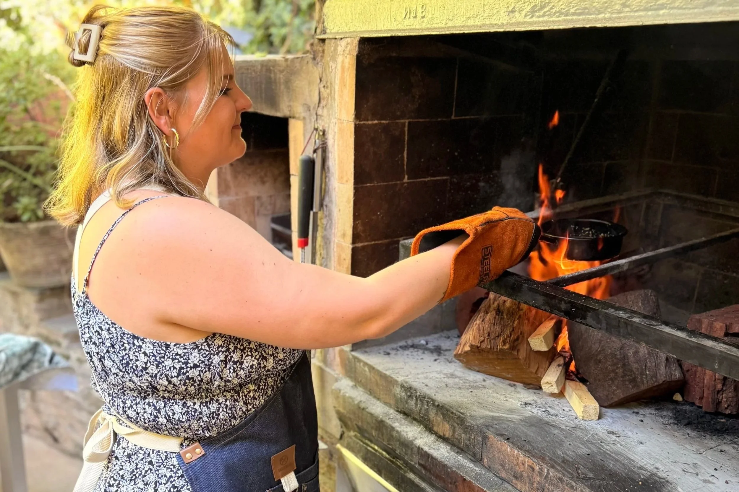 Piper wearing an apron and grill glove, tending a wood fire in a traditional brick parrilla.