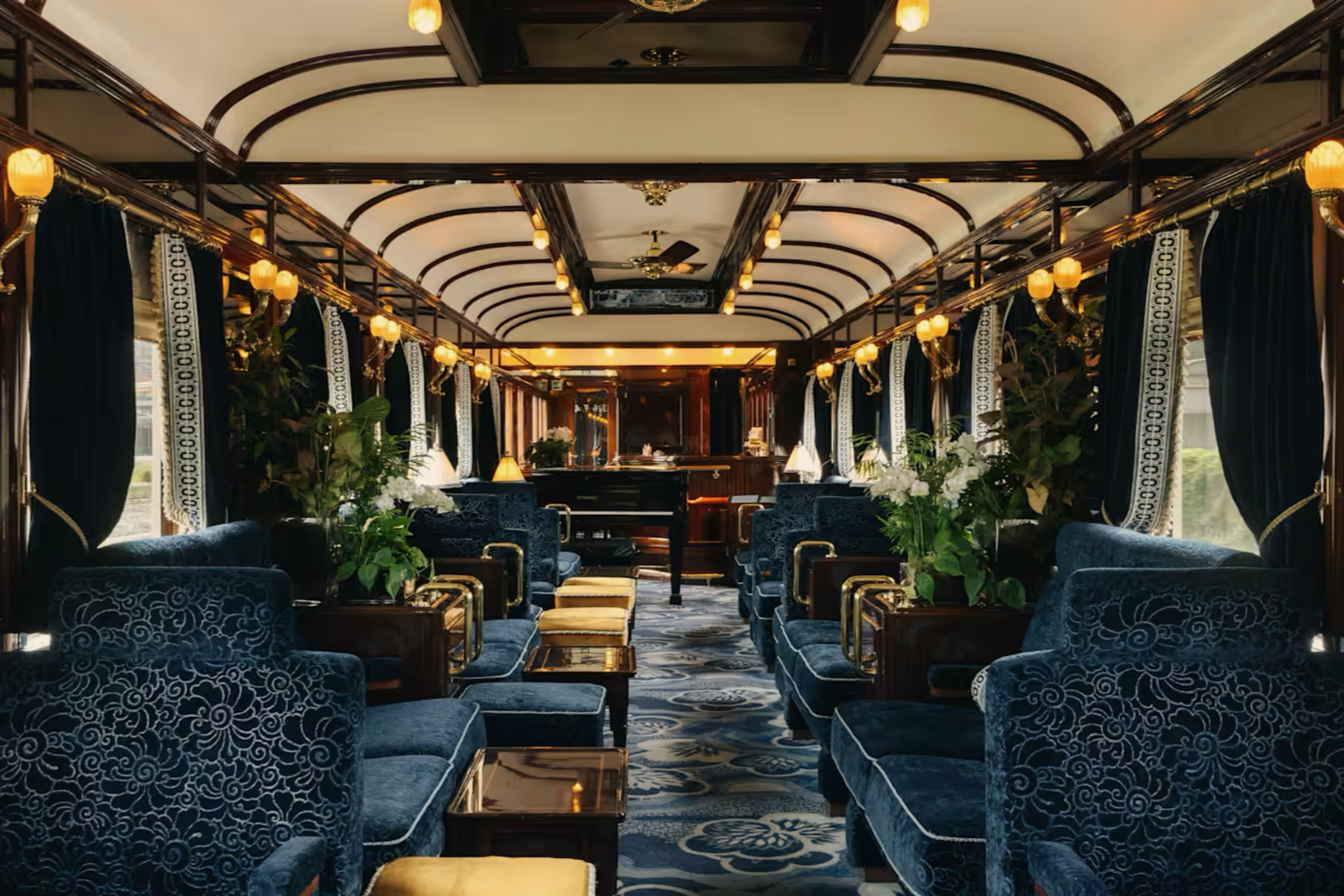 Interior of a vintage train car with blue velvet armchairs, gold accents, green plants, large windows with blue curtains, and ornate lighting fixtures.