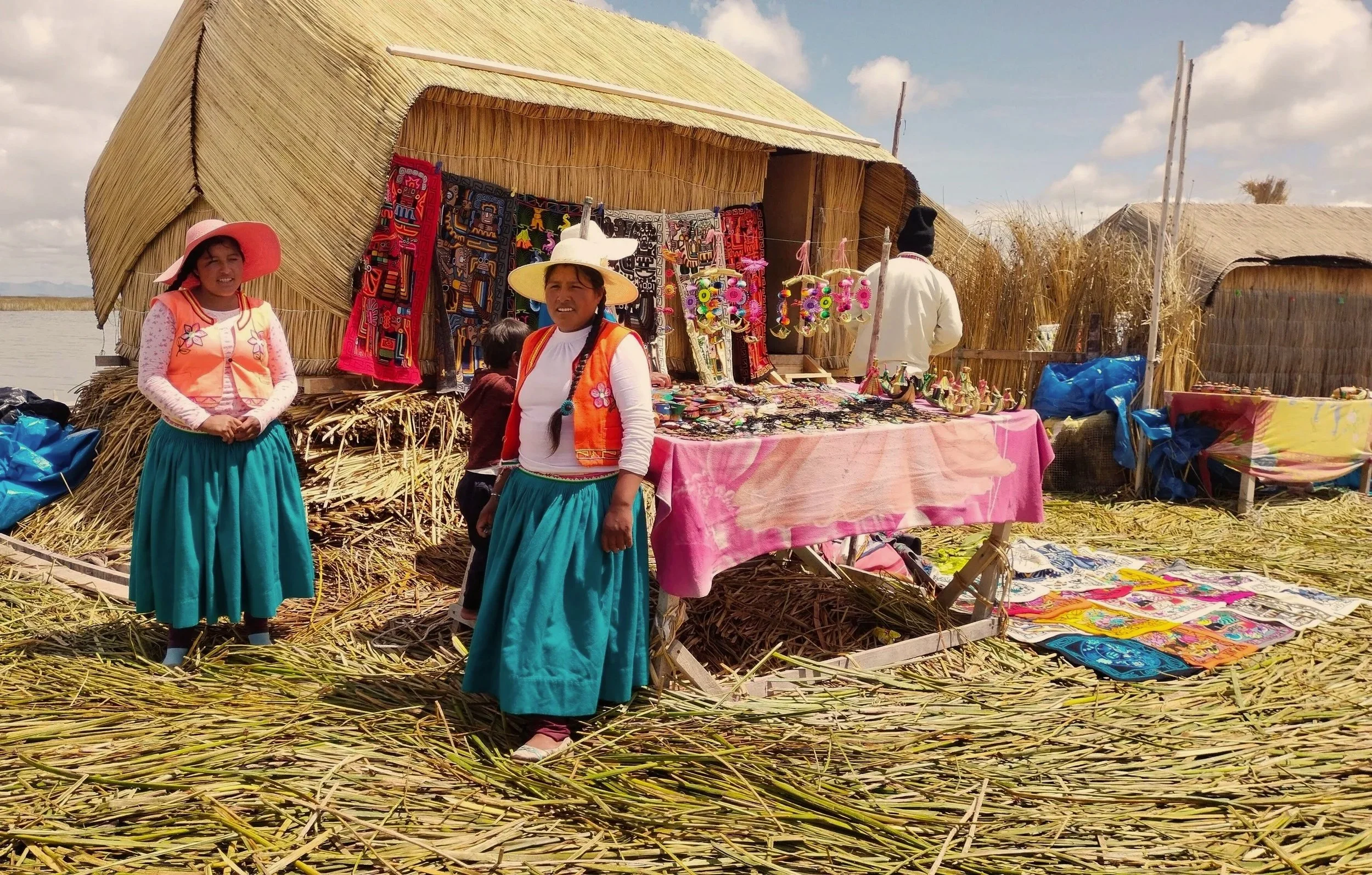 Two Uros women in traditional skirts and hats stand beside a totora reed hut and handmade textiles on a floating island in Lake Titicaca