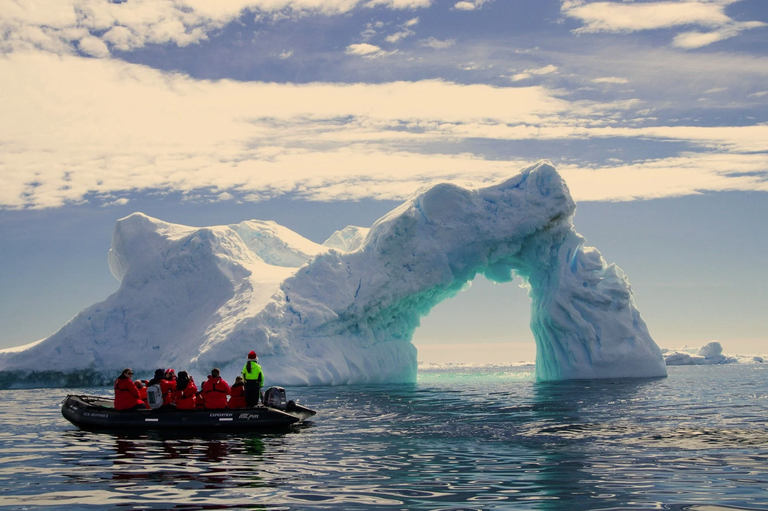 Group of people in red jackets on a boat observing a large iceberg in icy waters.