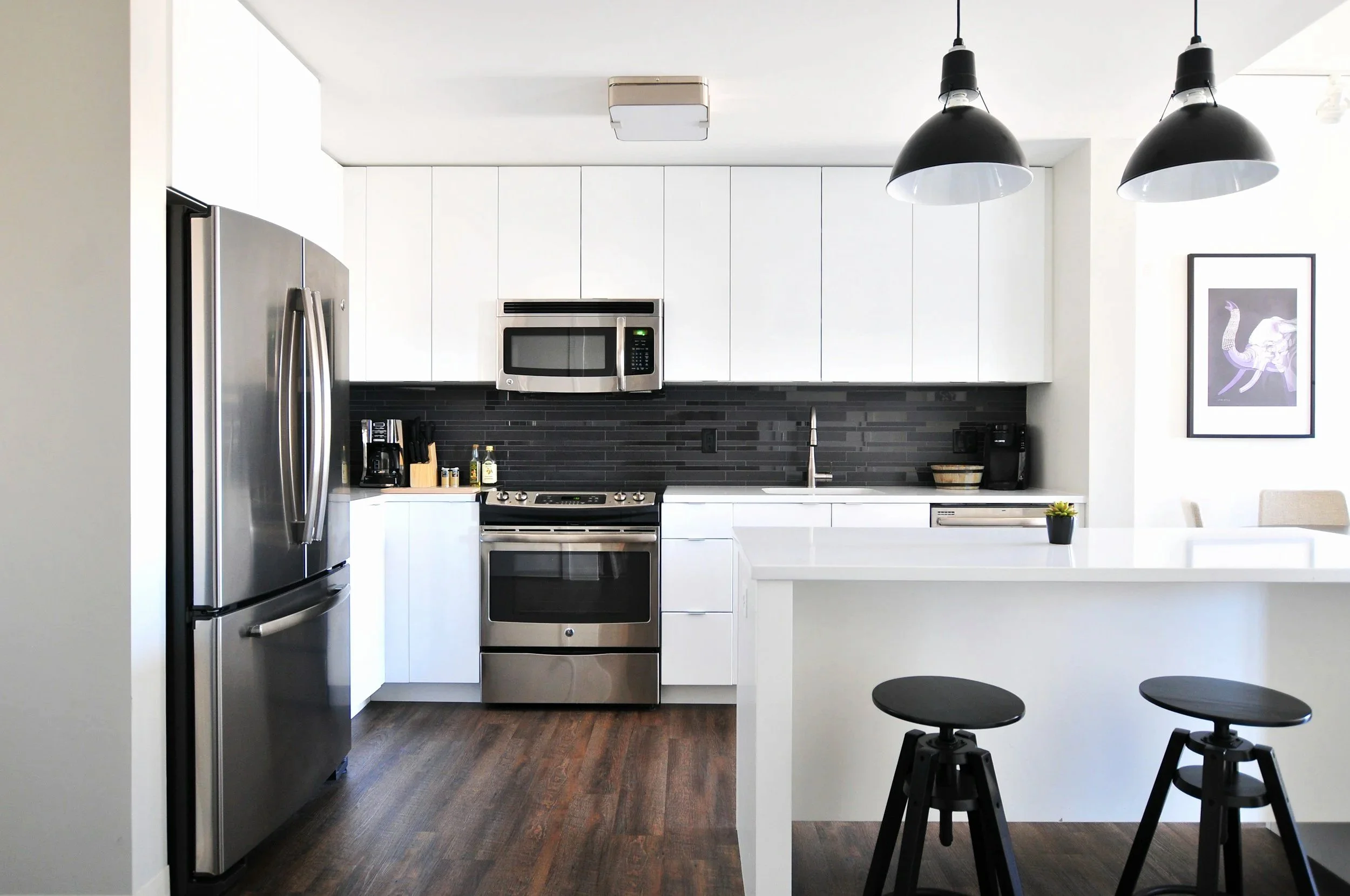 Modern kitchen with white cabinets, black backsplash, stainless steel appliances including a refrigerator, microwave, and oven, and black pendant lights above a white counter island. There are dark wood floors and minimalist bar stools around the island, with a framed artwork on the wall.