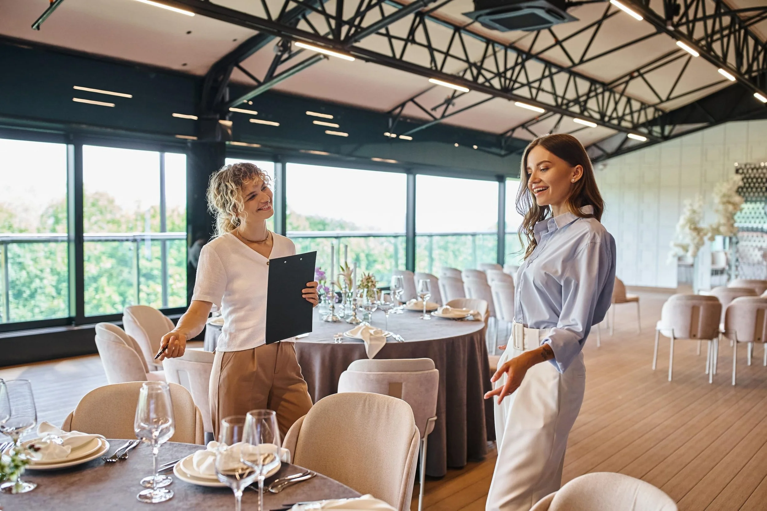 Two women having a conversation inside a stylish restaurant with large glass windows, decorated tables, and modern ceiling fixtures.