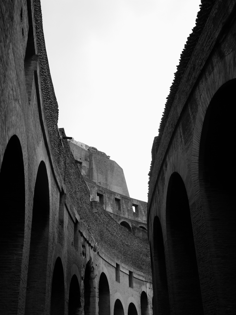 Black and white photo of the inside of the Colosseum in Rome, showing its arches and ancient stone structure from a low angle.