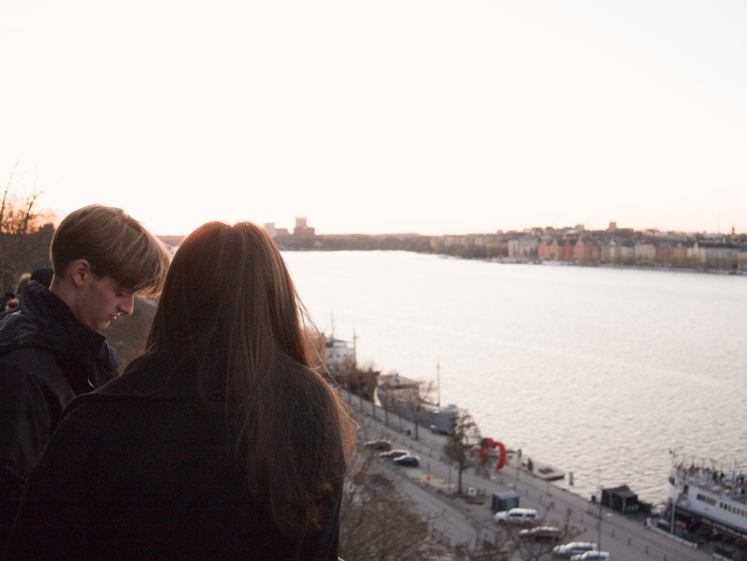 Two people with dark hair, one male and one female, standing near a river at sunset.