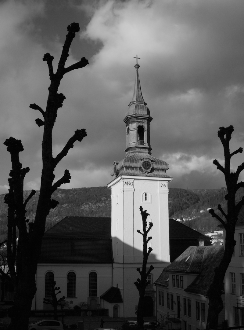 Black and white photo of a church with a tall steeple topped with a cross, surrounded by pruned trees with no leaves and a cloudy sky in the background.