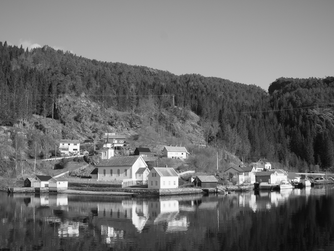A black and white photo of a small harbor town with houses and a church along the water, surrounded by forested hills.