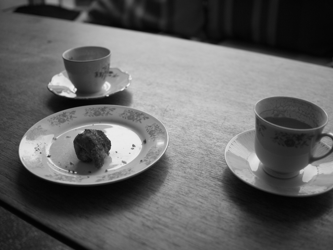 Two cups of tea or coffee on saucers and a partially eaten piece of cake or bread on a floral plate on a wooden table.