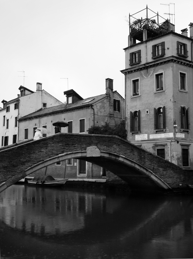 Black and white photo of a canal with a small arching bridge. A boat is docked under the bridge, and buildings with windows and chimneys line the canal. A person with an umbrella stands on the bridge.