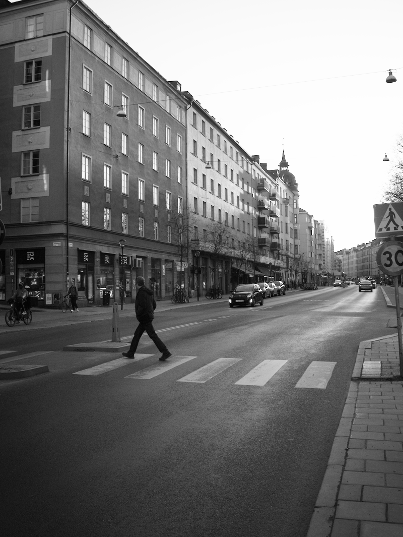 Black and white photo of a city street with a person walking across a crosswalk, multi-story buildings along the sidewalk, cars parked and driving, and a street sign indicating a 30 km/h speed limit.