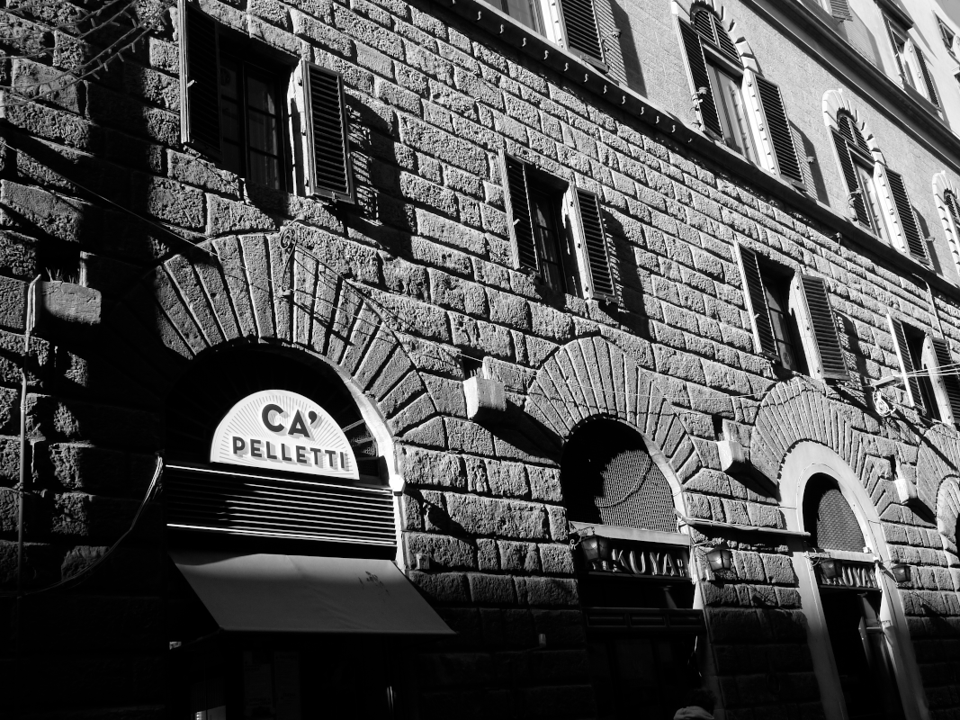 Black and white photo of an old brick building with arched windows and shutters, with signs for Ca' Pelletti and a storefront.