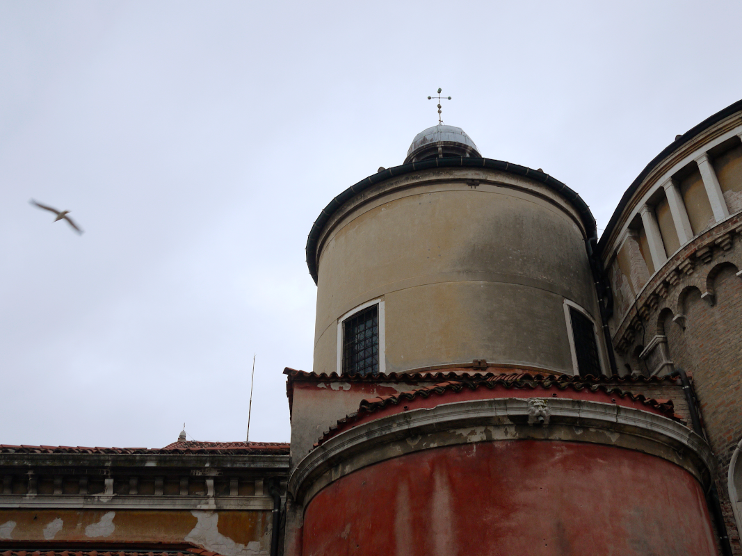 An old historic building with a rounded tower, weathered yellow and red walls, and a small window, with a cloudy sky and a bird flying nearby.