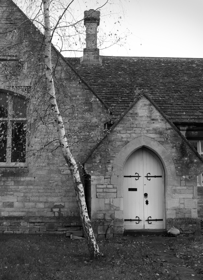 Black-and-white photo of an old stone house with a gabled roof, a chimney, and an arched white wooden door with decorative black hardware, next to a leafless tree in the yard.