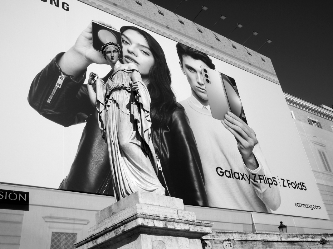 Large billboard advertising Samsung Galaxy Z Flip5 and Z Fold5 smartphones with images of a woman and a man holding the phones. In front of the billboard is a classical statue of a woman.
