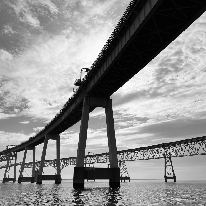 Black and white photo of a large bridge extending over water with a cloudy sky in the background.