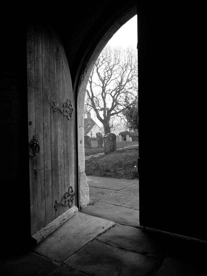 Open wooden church door revealing a graveyard with leafless trees and headstones outside on a cloudy day.