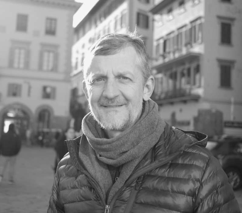 A middle-aged man with a beard, smiling, wearing a puffer jacket and scarf, outdoors on a city street with buildings and people in the background.