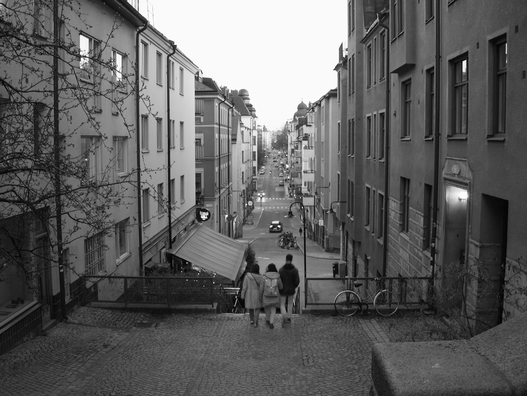 A black and white photo of a city street viewed from a set of stairs. Three people walk down the stairs towards the street, which has cars, bicycles, and buildings on both sides.