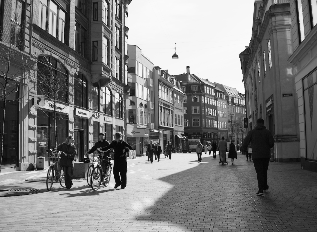 Street scene with people walking and two people with bicycles on a sunny day in an urban area with multi-story buildings.