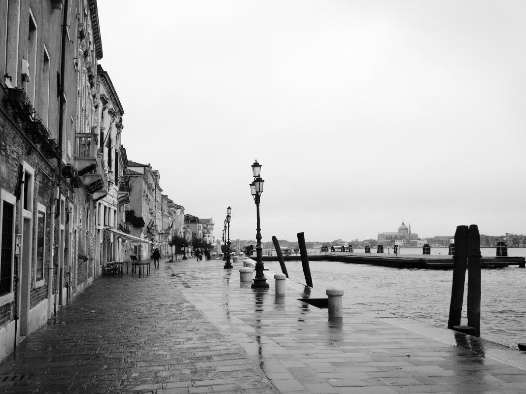A black and white photo of a riverside walkway with old buildings on the left, street lamps, a few pedestrians, and a view of a building with a dome in the distance on the right.