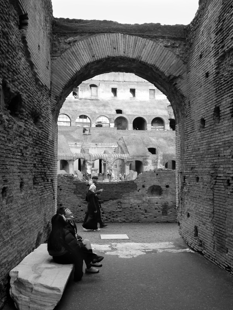 View inside the ancient Roman Colosseum showing archways and tiered seating areas with tourists. Some visitors are sitting and some are walking.