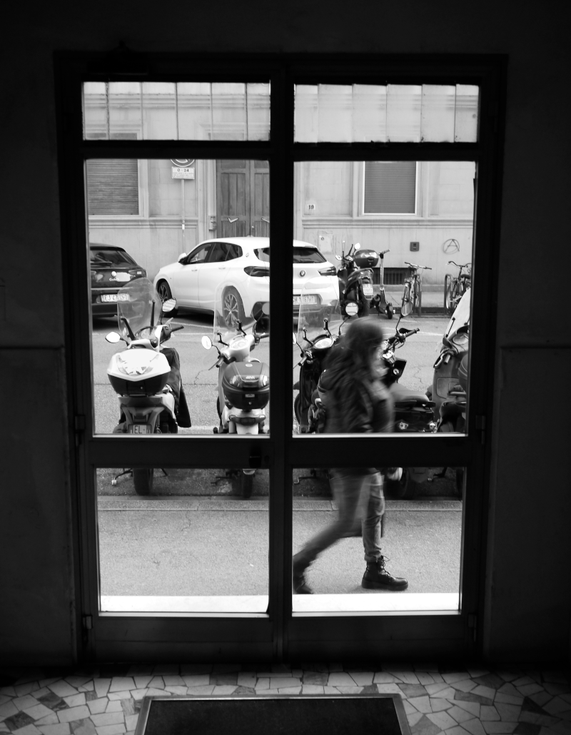 A person walking past a window with a view of parked motorcycles and cars on the street outside.