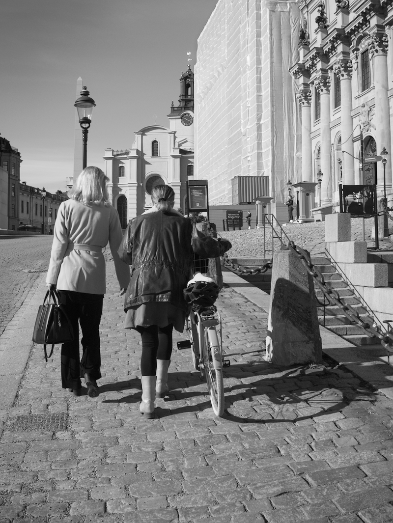 Two women walking on cobblestone street, one pushing bicycle, near historic building with scaffolding and architectural details, under clear sky.