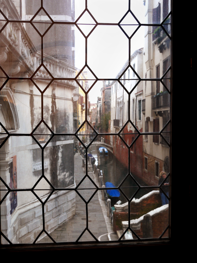 View of a narrow canal street in Venice, Italy, seen through an ornate iron lattice window, with buildings on either side and a small boat in the canal.