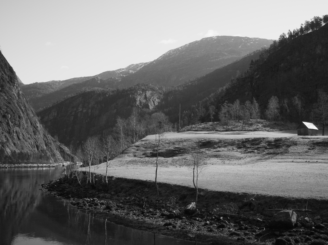 Scenic black and white landscape of a lake, mountains, and a small house in a rural area.