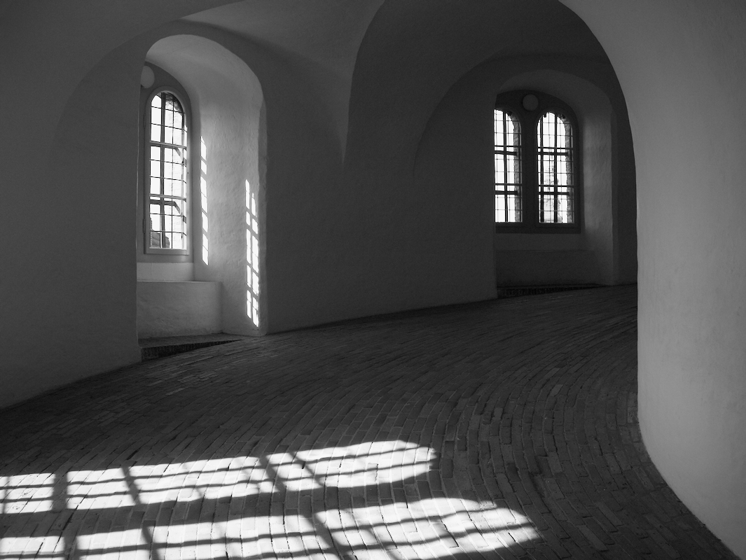 Black and white photo of an interior space with arched windows and sunlight casting shadows onto the brick floor.