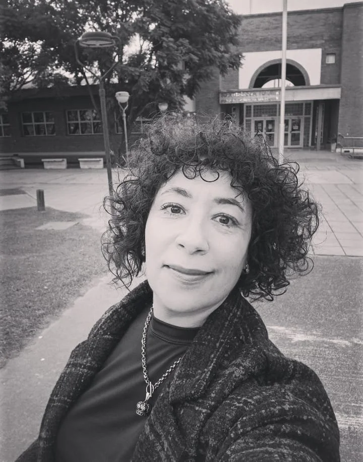 Retrato en blanco y negro de una mujer con cabello rizado y oscuro, sonriendo ligeramente en una calle frente a un edificio. Ella lleva una bufanda o abrigo y un collar, con árboles y farolas en el fondo.