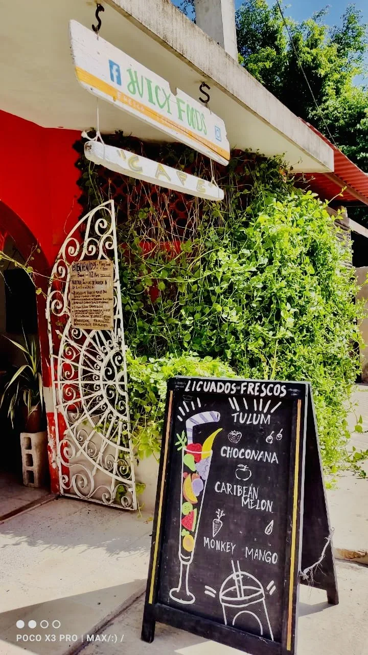 A chalkboard sign outside a juice bar displays various blended fruit drinks, including Tulum, Choconana, Caribbean Melon, Monkey Mango, with drawings of a tall glass and fruit.