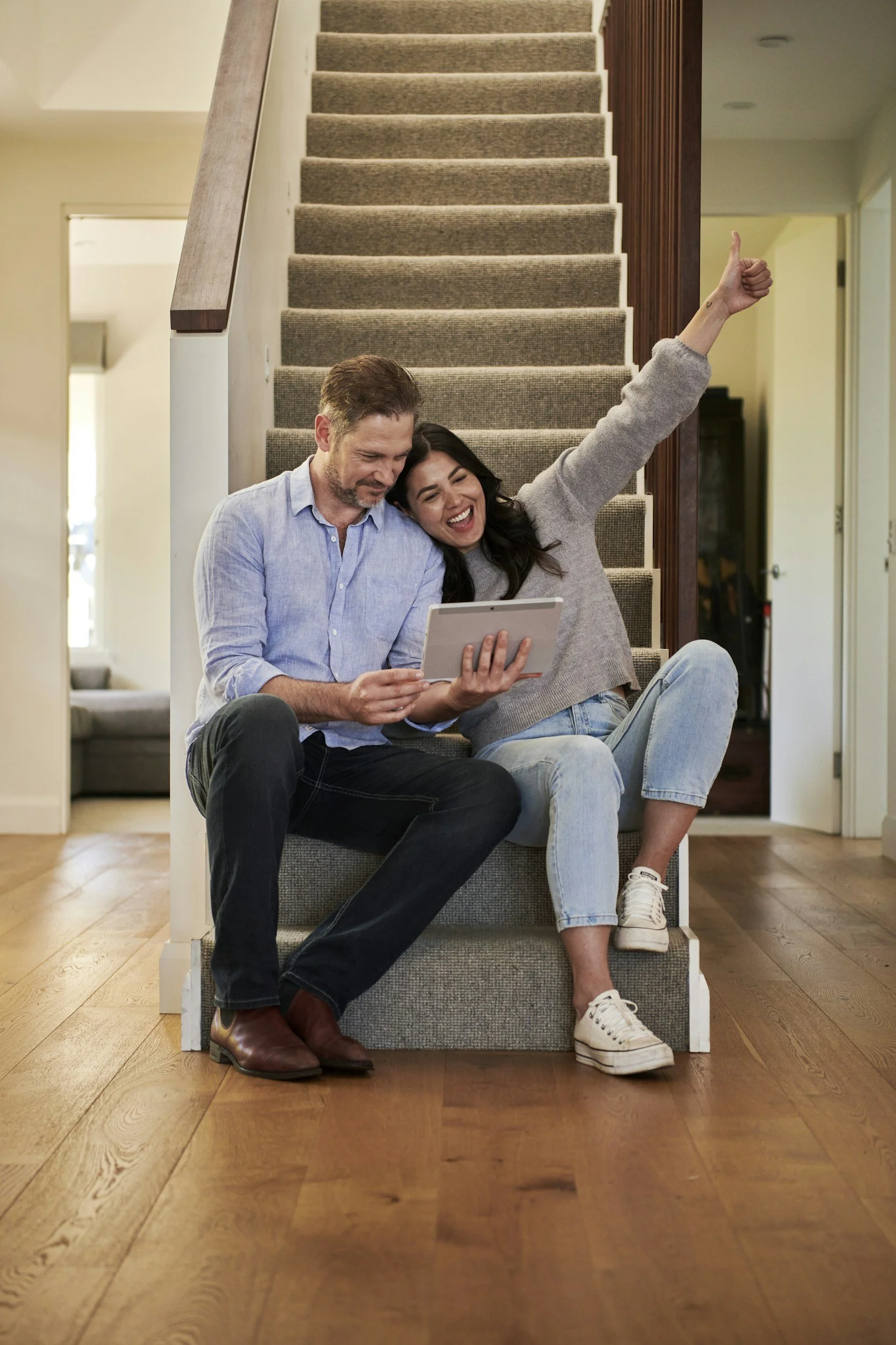 A couple sitting on stairs in a home, looking at a tablet. The woman is giving a thumbs-up while smiling, and the man is looking at the tablet with a slight smile. The stairs are carpeted, and the setting appears cozy and modern.