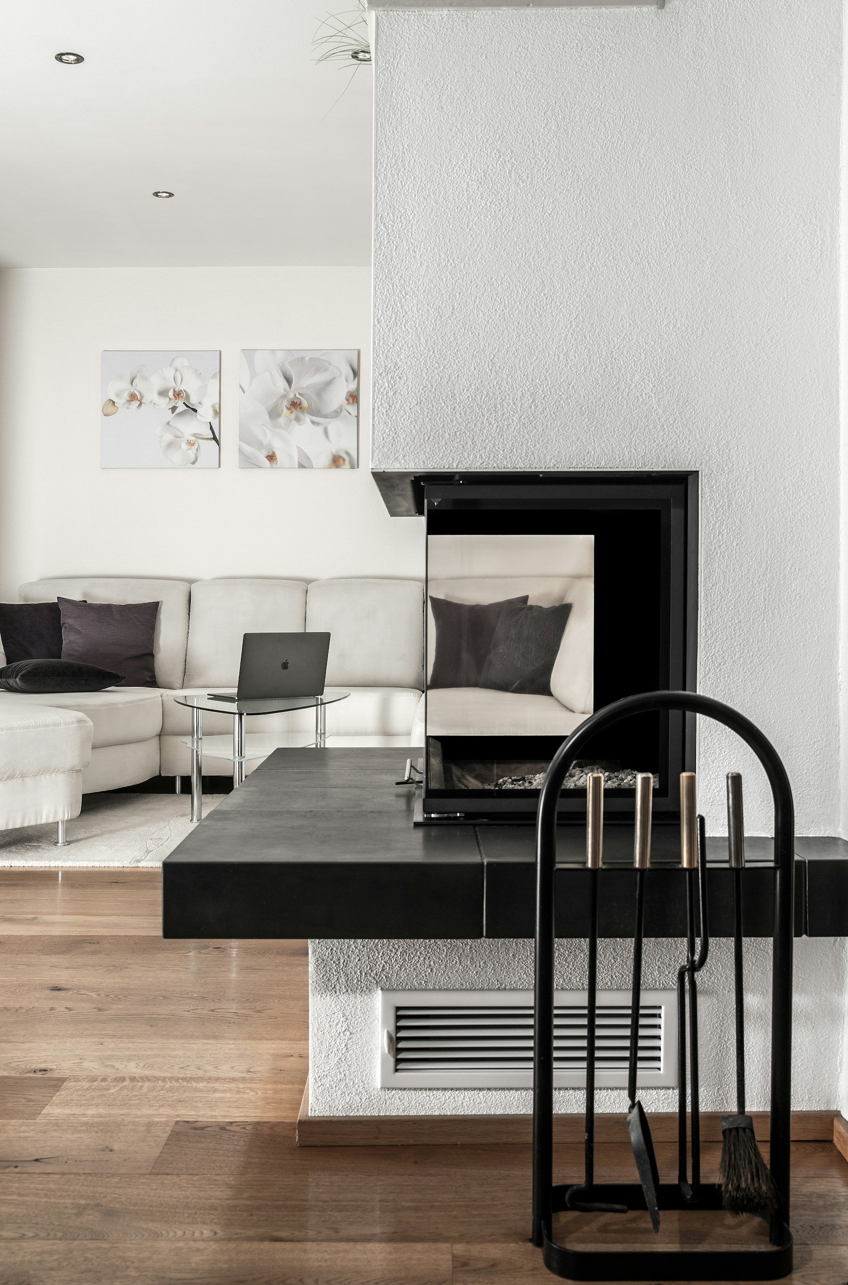 Living room with white couch, black pillows, abstract floral wall art, fireplace, laptop on table, hardwood floor, and black fireplace tools in the foreground.