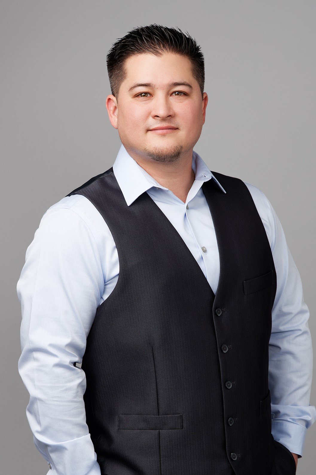Professional headshot of a man with short dark hair, wearing a white dress shirt and black vest, standing against a plain gray background.