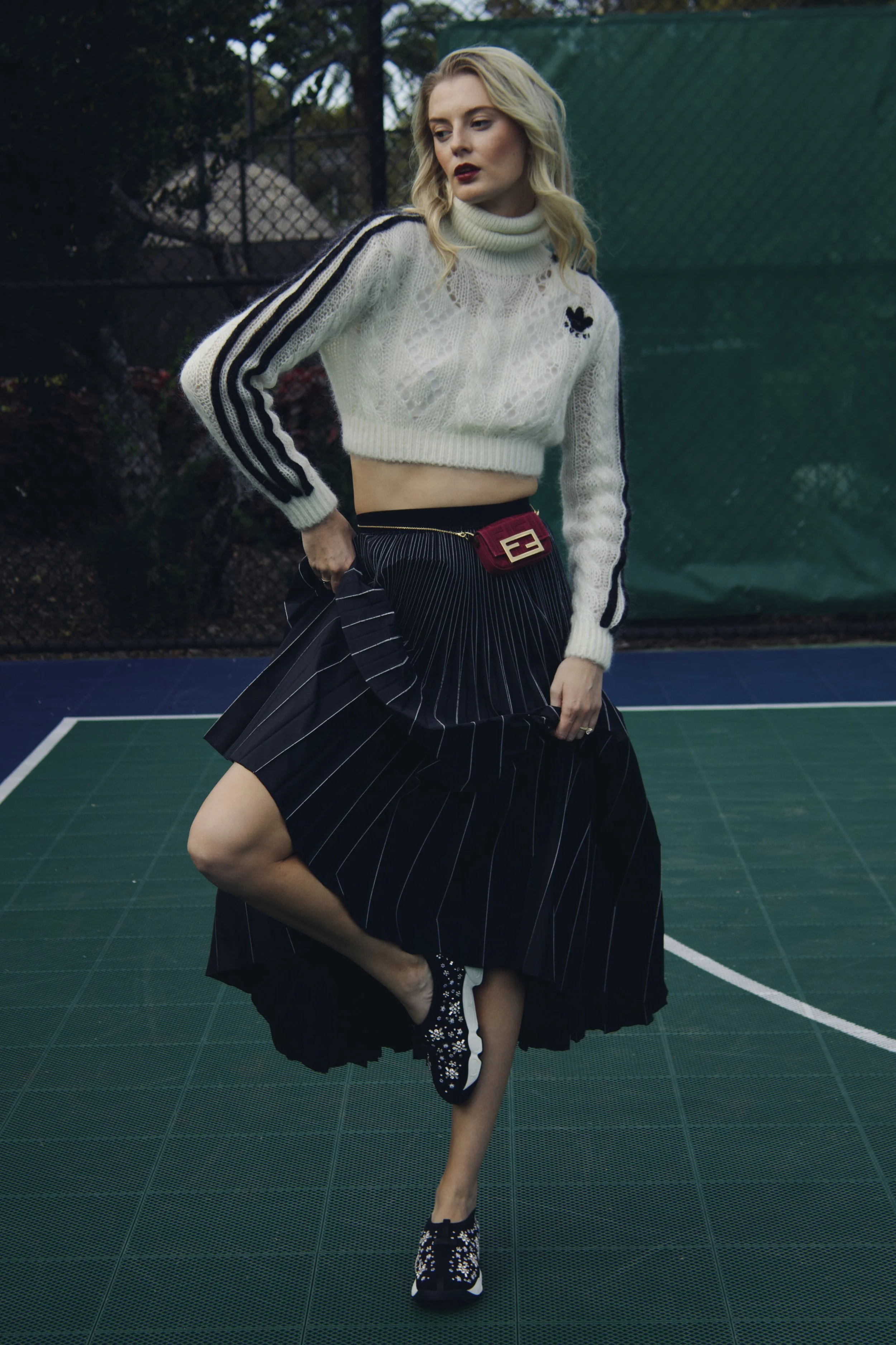 a model stands on a basketball court wearing a gucci sweater, black pleated skirt, fendi mini bag, and dior sneakers