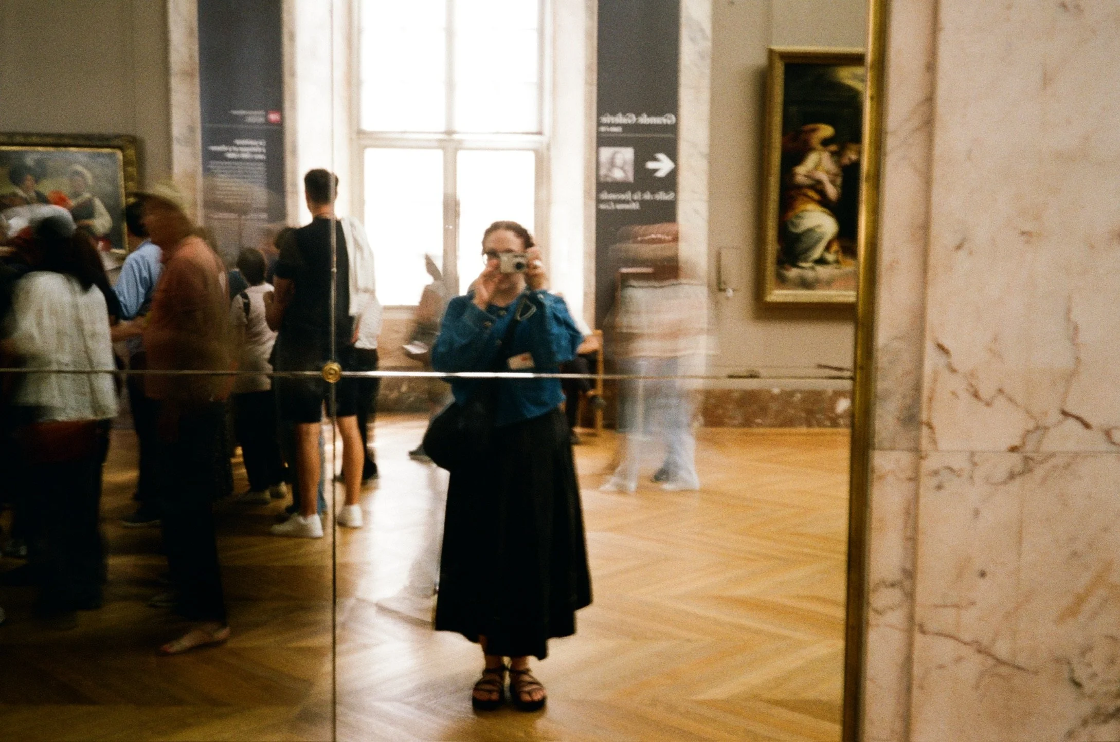 A woman taking a photo of herself in a large museum hallway with other visitors, paintings on the walls, a large window, and a marble column on the right.
