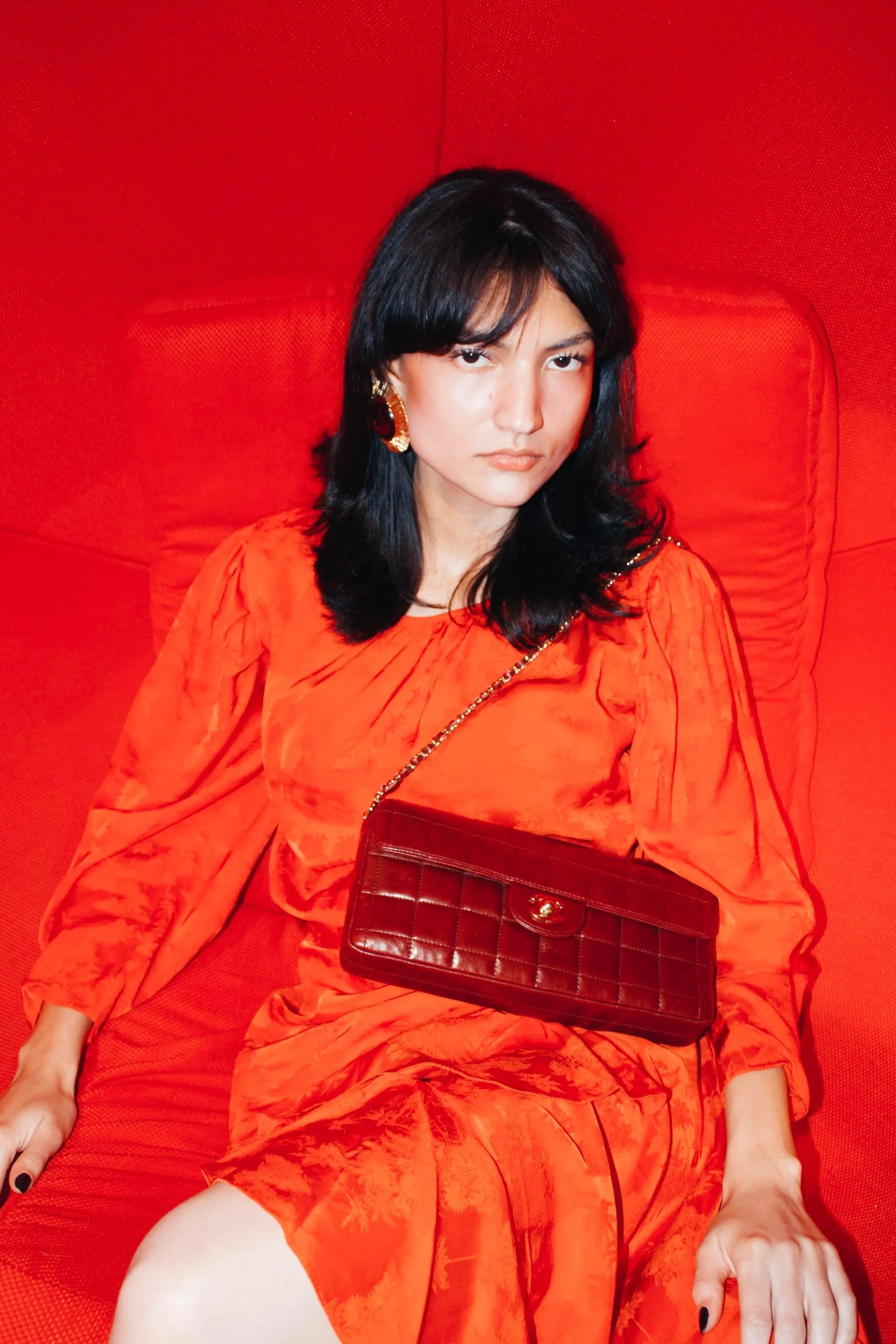 a model sits in a red egg chair wearing a johanna ortiz red dress and a chanel burgundy vintage chocolate bar bag