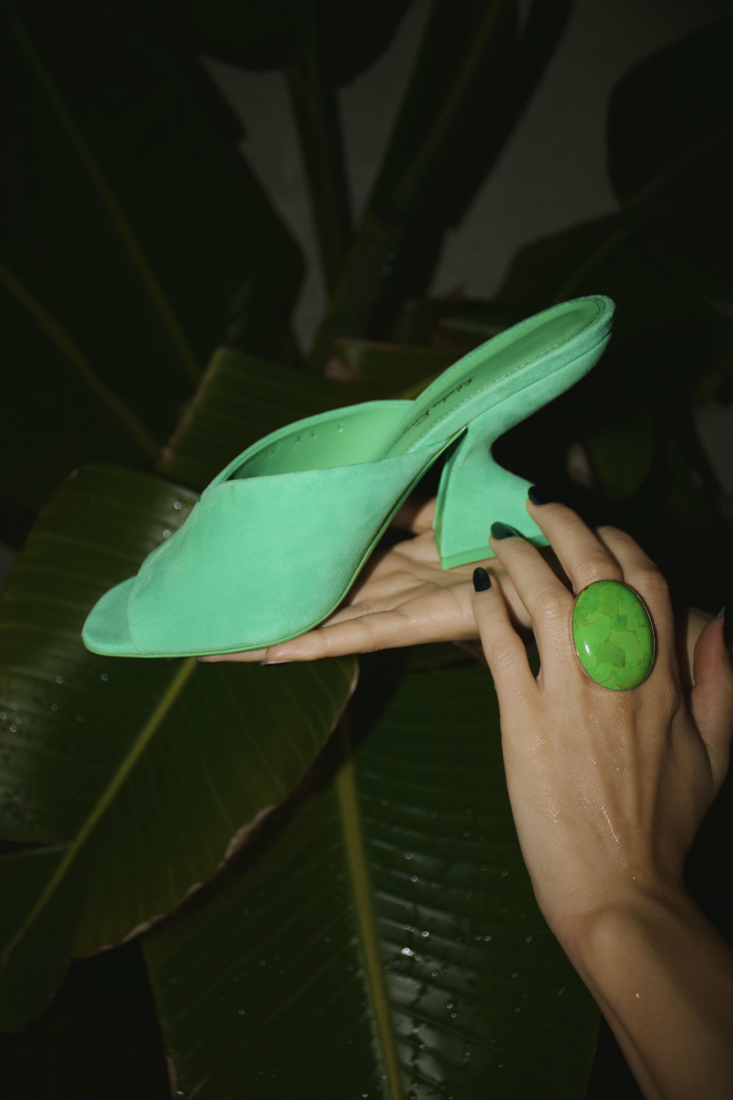 a flash photograph of a hand holding a Ferragamo green suede sculpted heel mule in front of palm trees while wearing a green stone ring