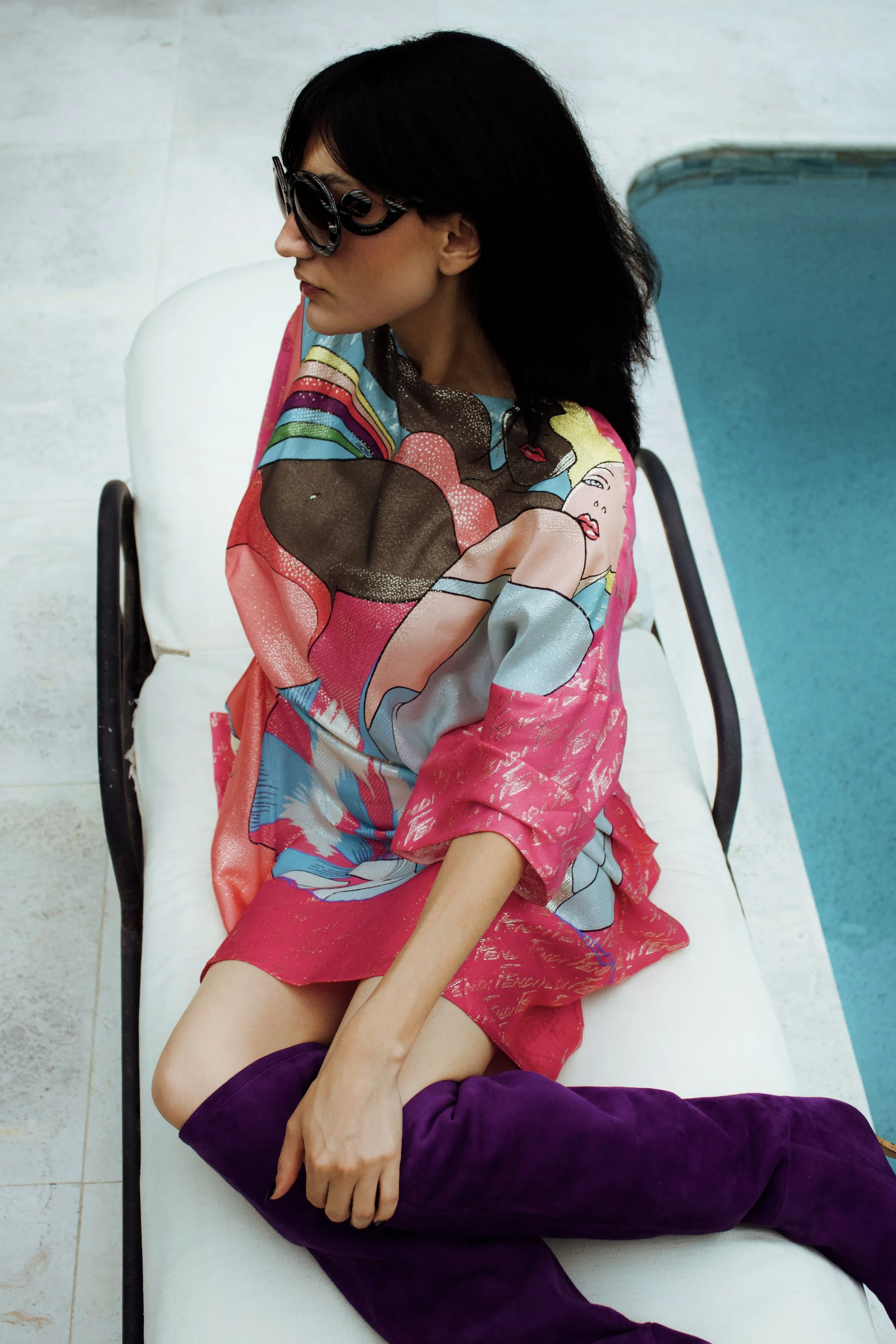 model in a fendi pink graphic print dress and purple knee high boots sitting on a deck chair beside a pool wearing prada round sunglasses
