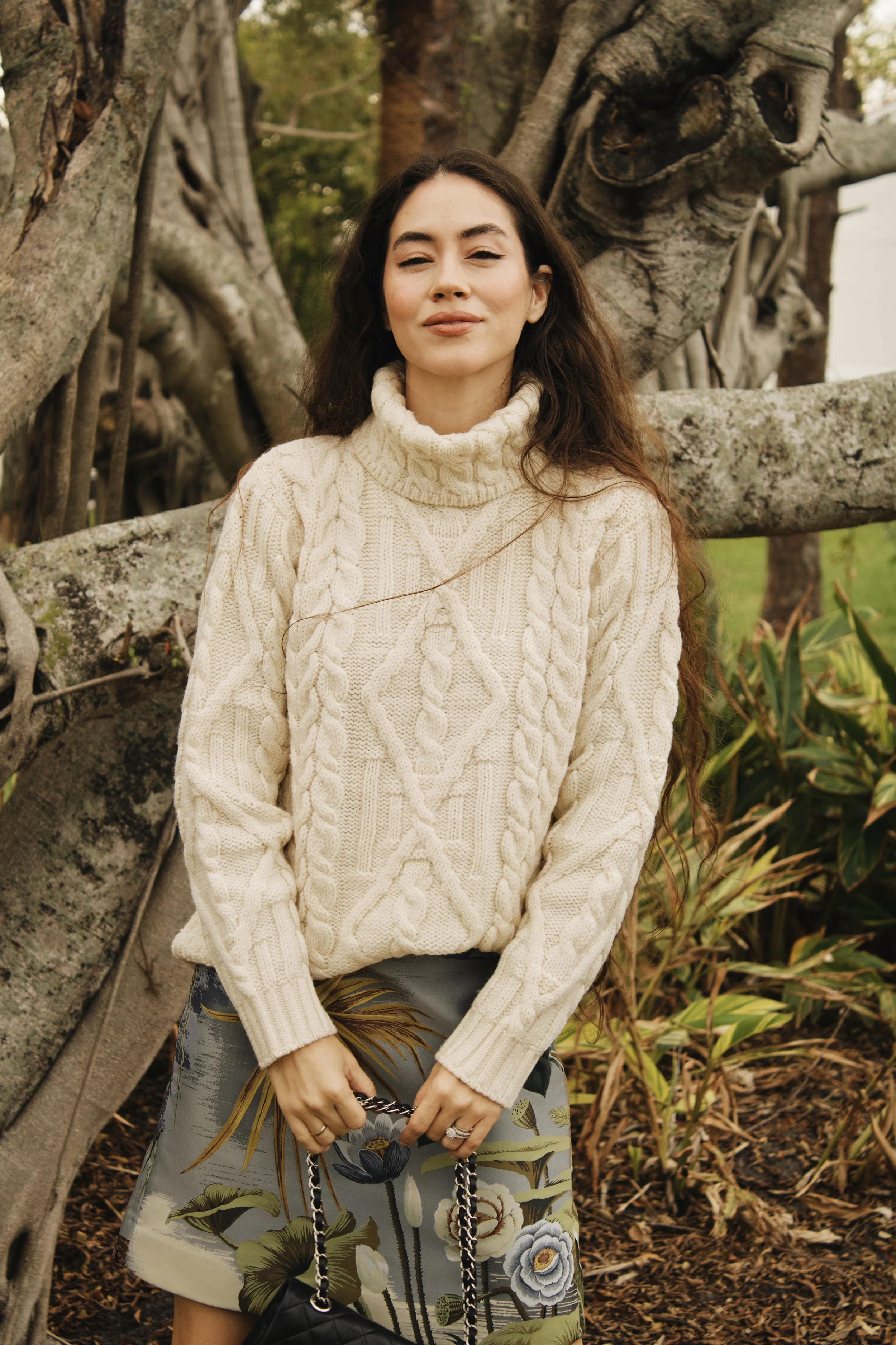 model in a cable knit sweater underneath trees and plants in the fall