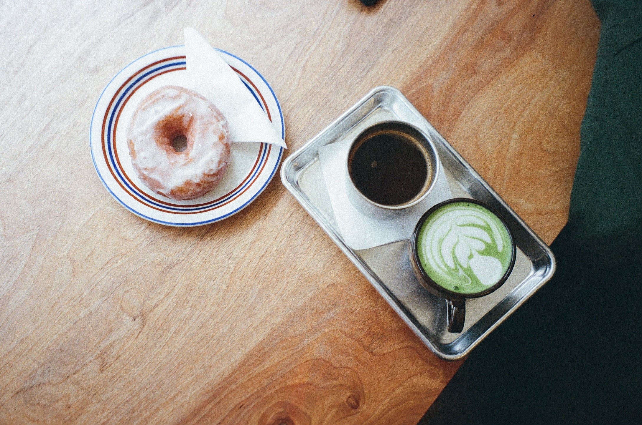 A wooden table with a plate holding a glazed donut on a paper doily, a silver tray with a cup of black coffee, and a cup of green matcha latte with foam art.