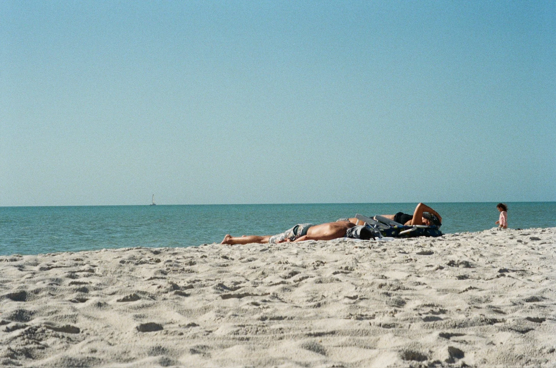 People relaxing on a sandy beach near the ocean with a sailboat in the distance under a clear blue sky.