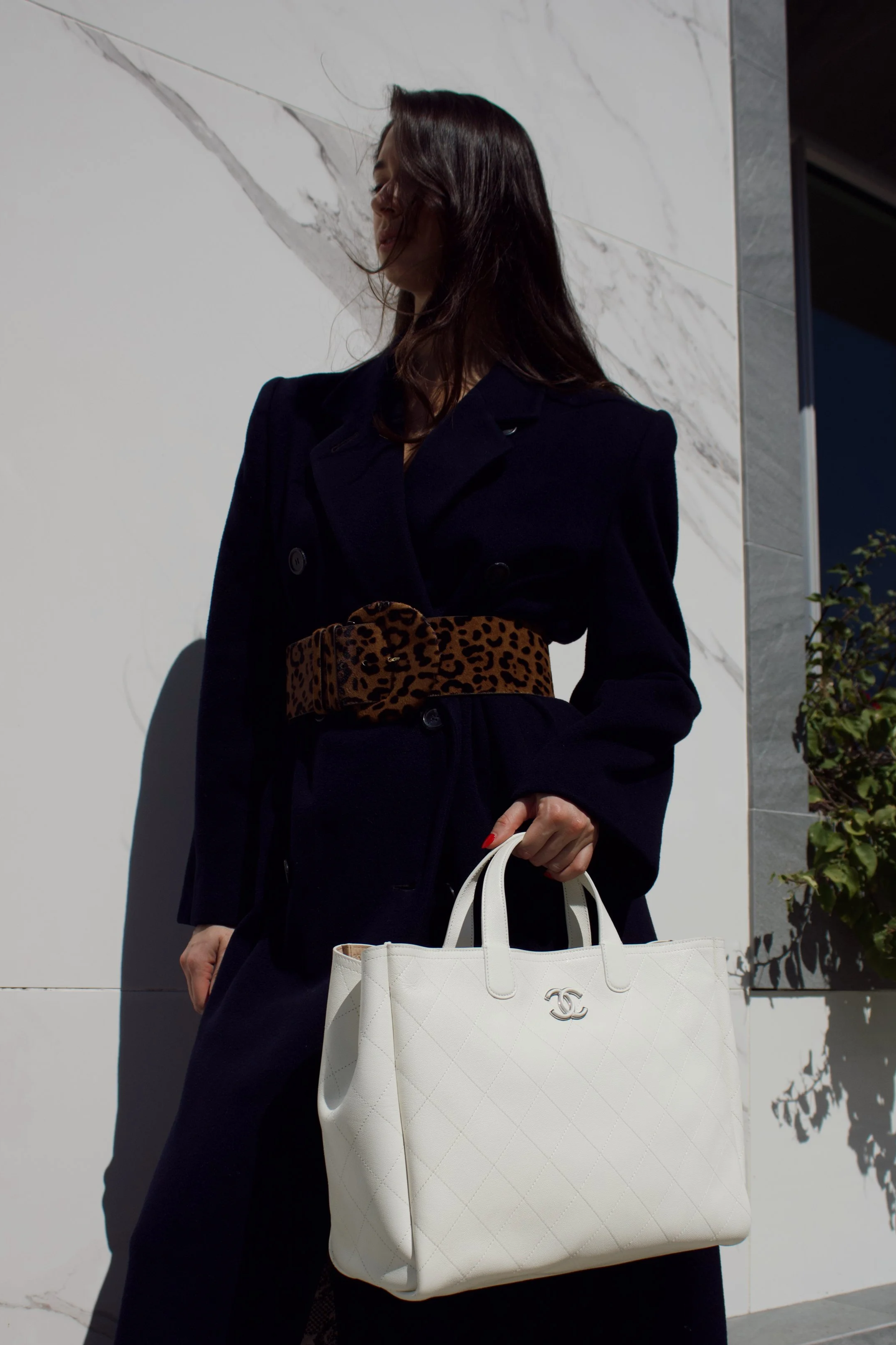 a model wears a long black coat and holds a chanel white leather tote while standing in front of a marble wall