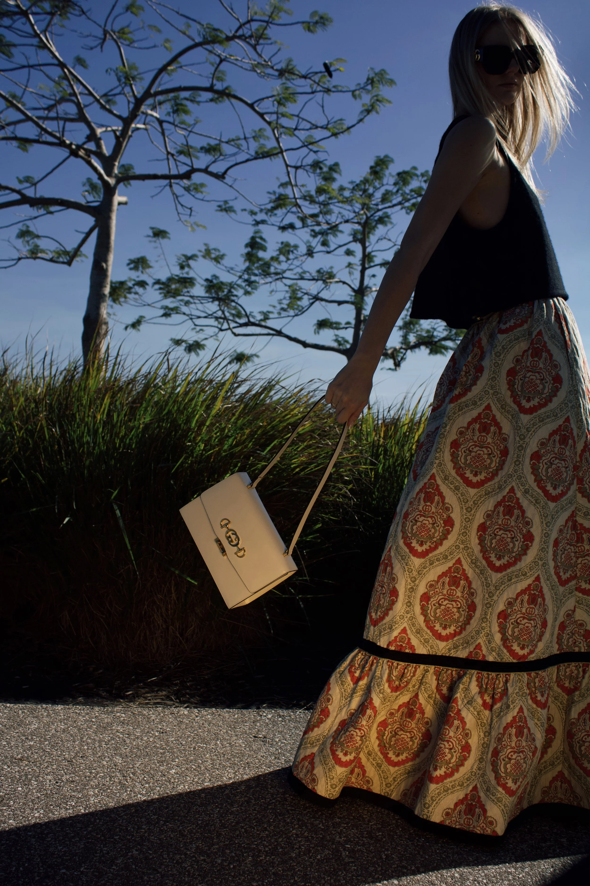 a model walks by wearing a vintage gucci long skirt swinging a white gucci bag with a tree and grass in the background