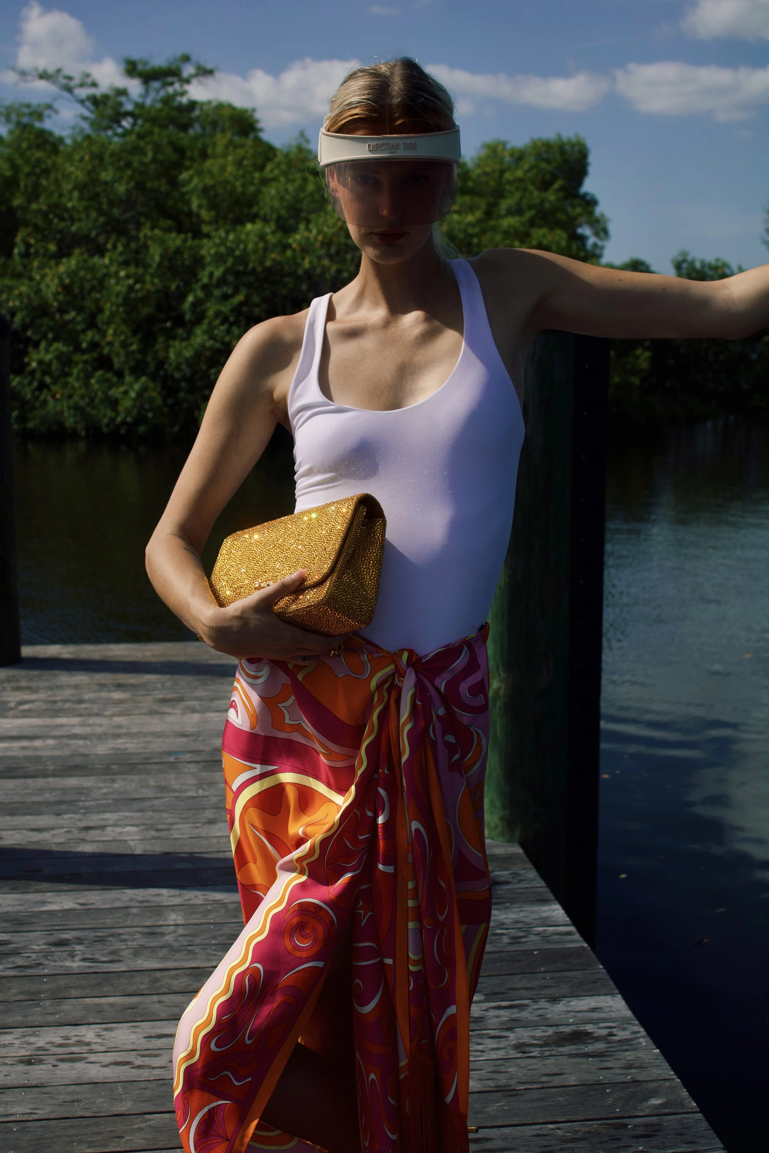 a model leans on a pier wearing a dior visor, pucci orange skirt, and white body suit while holding a gold bedazzled chanel bag