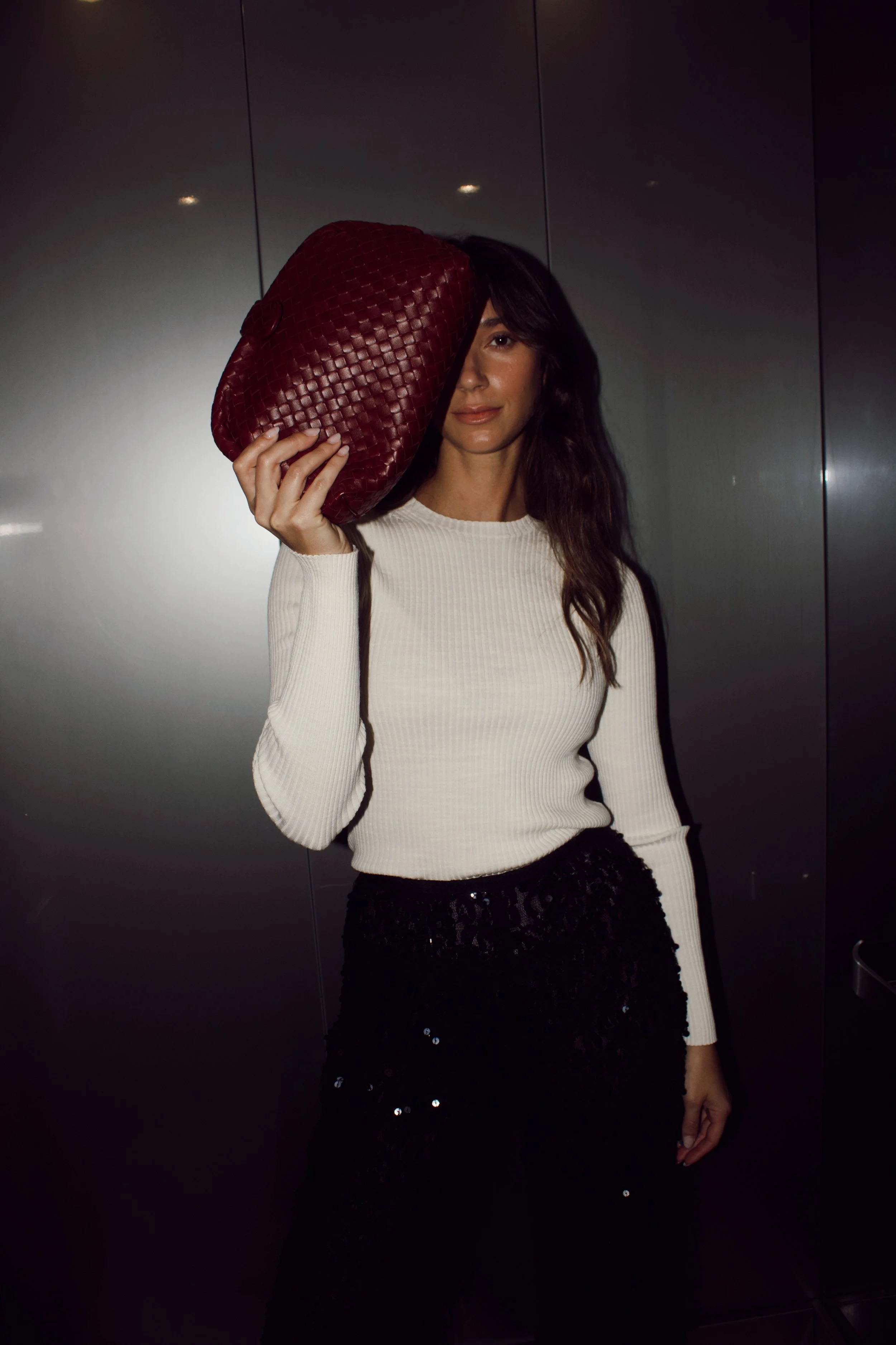 a flash photograph of a model standing in an elevator holding a burgundy bottega veneta intrecciato clutch over her face