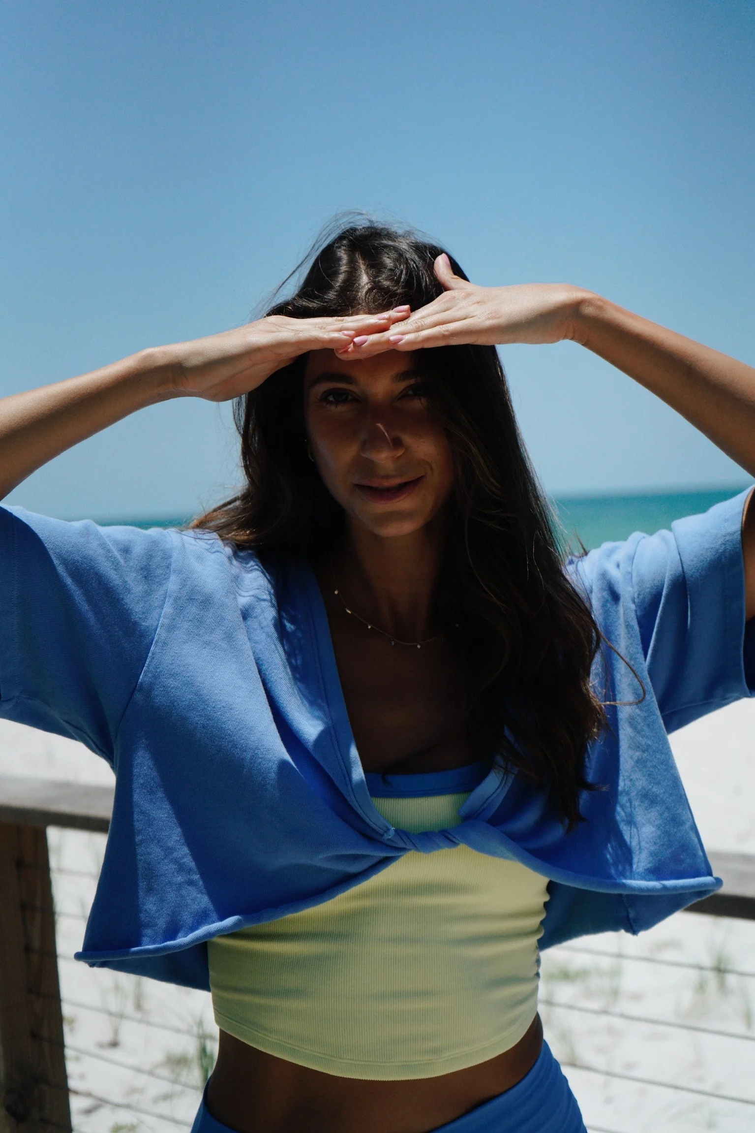 Young woman on the beach wearing a blue jacket, yellow top, and blue pants, with her hands on her forehead, looking into the distance.